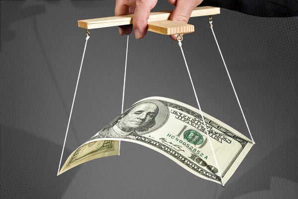 A photo illustration of a 100-dollar bill puppet on strings being manipulated by a hand.