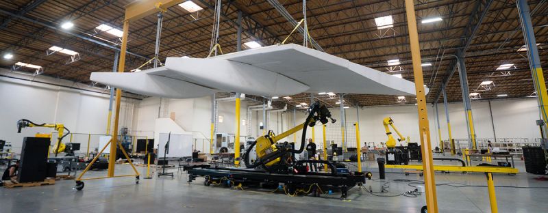 A white aircraft wing suspended above a mobile robot with a large yellow arm in an open hangar space.